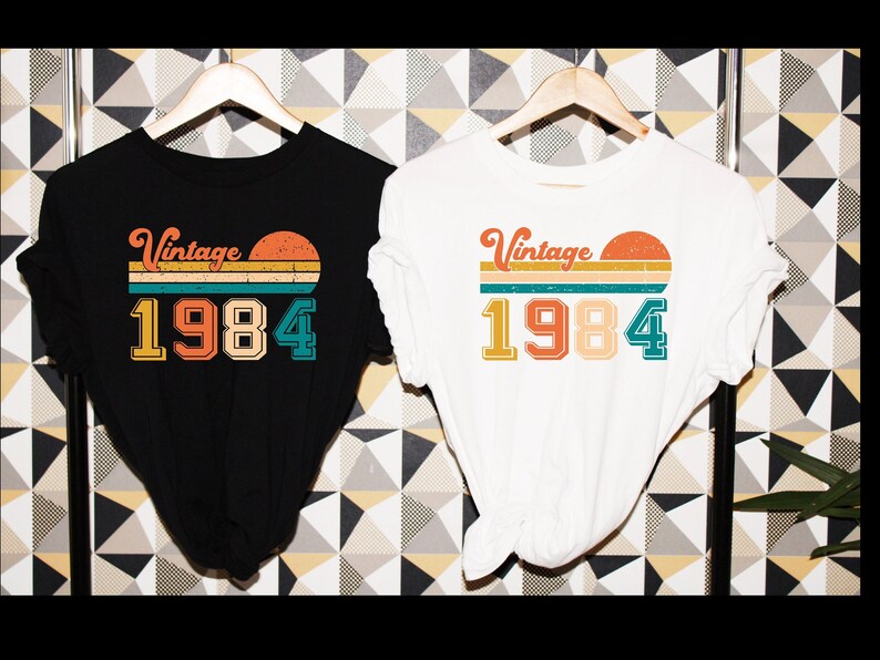 Peut inclure: Deux t-shirts avec un design vintage 1984. Le t-shirt noir a du texte blanc et le t-shirt blanc a du texte noir. Les deux t-shirts pr&eacute;sentent un graphique de coucher de soleil avec un design ray&eacute;.