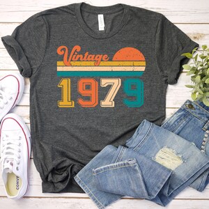 Może przedstawiać: Szary t-shirt z nadrukiem w stylu vintage z rokiem 1979 i zachodem słońca. Grafika jest w odcieniach pomarańczowego, ż&oacute;łtego, zielonego i niebieskiego.