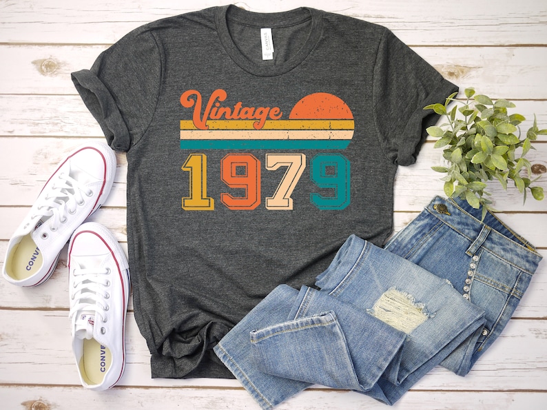 Może przedstawiać: Szary t-shirt z nadrukiem w stylu vintage z rokiem 1979 i zachodem słońca. Grafika jest w odcieniach pomarańczowego, ż&oacute;łtego, zielonego i niebieskiego.