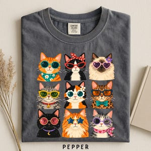 A gray t-shirt with a graphic design of nine cats wearing sunglasses and accessories, arranged in a 3x3 grid.