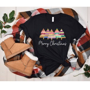 K&ouml;nnte beinhalten: Schwarzes T-Shirt mit einem Motiv von drei bunten Weihnachtsb&auml;umen und dem Text "Frohe Weihnachten". Das Shirt liegt auf einer karierten Decke mit braunen Wildlederschuhen.