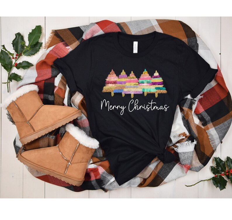 K&ouml;nnte beinhalten: Schwarzes T-Shirt mit einem Motiv von drei bunten Weihnachtsb&auml;umen und dem Text "Frohe Weihnachten". Das Shirt liegt auf einer karierten Decke mit braunen Wildlederschuhen.
