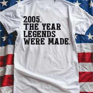 A white t-shirt with the text "2005. THE YEAR LEGENDS WERE MADE." printed on it, placed on top of an American flag.