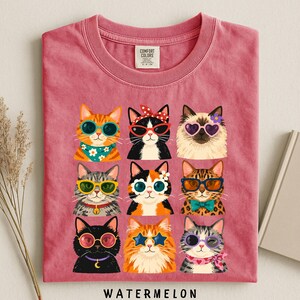 A pink t-shirt with a collage of nine cat illustrations wearing sunglasses and accessories, arranged in a 3x3 grid.