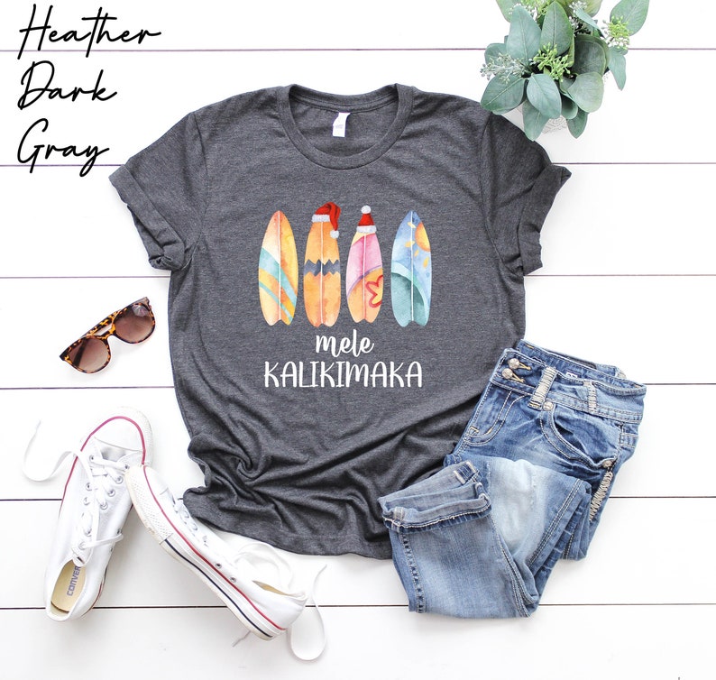 Op de afbeelding: Heather donkergrijs T-shirt met een aquarelontwerp van vier surfplanken waarvan er &eacute;&eacute;n een kerstmanmuts draagt. De tekst "mele kalikimaka" is onder de surfplanken gedrukt.