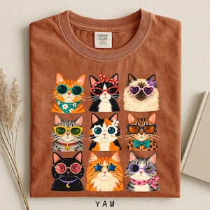 A t-shirt with a collage of nine cat illustrations wearing sunglasses and accessories, arranged in a 3x3 grid.