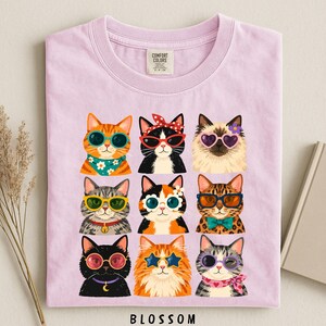 A pink t-shirt with a collage of nine cat illustrations wearing sunglasses and accessories, arranged in a 3x3 grid.
