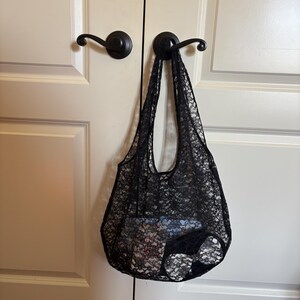 May include: A black lace tote bag hangs from a door handle. The bag is made of a delicate, open-weave lace fabric. The bag is filled with items, suggesting it's a reusable shopping bag or a stylish accessory.