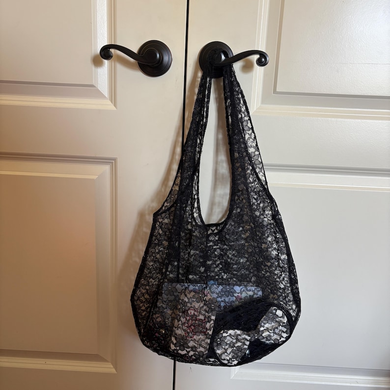 May include: A black lace tote bag hangs from a door handle. The bag is made of a delicate, open-weave lace fabric. The bag is filled with items, suggesting it's a reusable shopping bag or a stylish accessory.