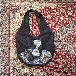 May include: Black lace tote bag with a matching wallet and card holder. The bag features a unique hourglass design in the center. The accessories are placed on a patterned rug.