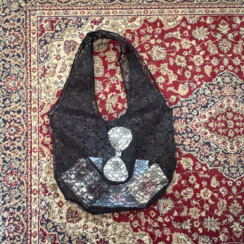 May include: Black lace tote bag with a matching wallet and card holder. The bag features a unique hourglass design in the center. The accessories are placed on a patterned rug.