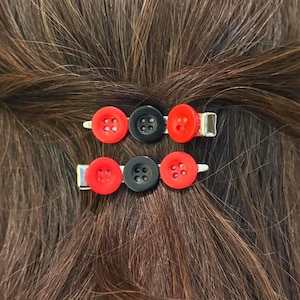 May include: Two hair clips, each featuring three circular buttons arranged in a row. The buttons are coloured red, black, and red, and each has four holes in the centre. The clips are silver-toned metal and are attached to dark brown hair.