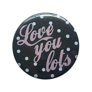 May include: A round button with a black background and white polka dots. The button features the phrase "Love you lots" in pink cursive lettering. The button is a simple, yet sweet, design.