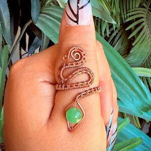 May include: A copper wire ring with a snake-like design, featuring a green gemstone. The ring wraps around the finger, with a coiled head and a body of woven wire. The gemstone is a round, light green bead.