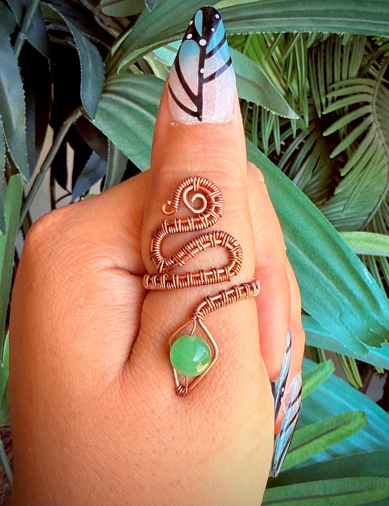 May include: A copper wire ring with a snake-like design, featuring a green gemstone. The ring wraps around the finger, with a coiled head and a body of woven wire. The gemstone is a round, light green bead.