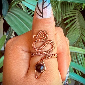May include: A copper wire ring in the shape of a snake, with a dark, round stone at the bottom. The ring is wrapped around a finger. The background is blurred green foliage.