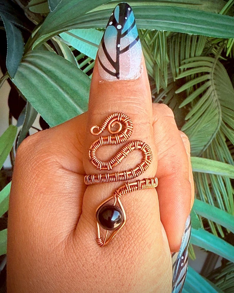 May include: A copper wire ring in the shape of a snake, with a dark, round stone at the bottom. The ring is wrapped around a finger. The background is blurred green foliage.