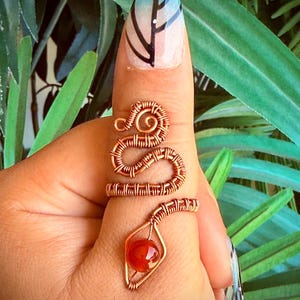 May include: A copper-colored wire-wrapped ring in the shape of a snake, with a red gemstone at the bottom. The ring is worn on a finger with a butterfly nail design. The snake's body coils around the finger.