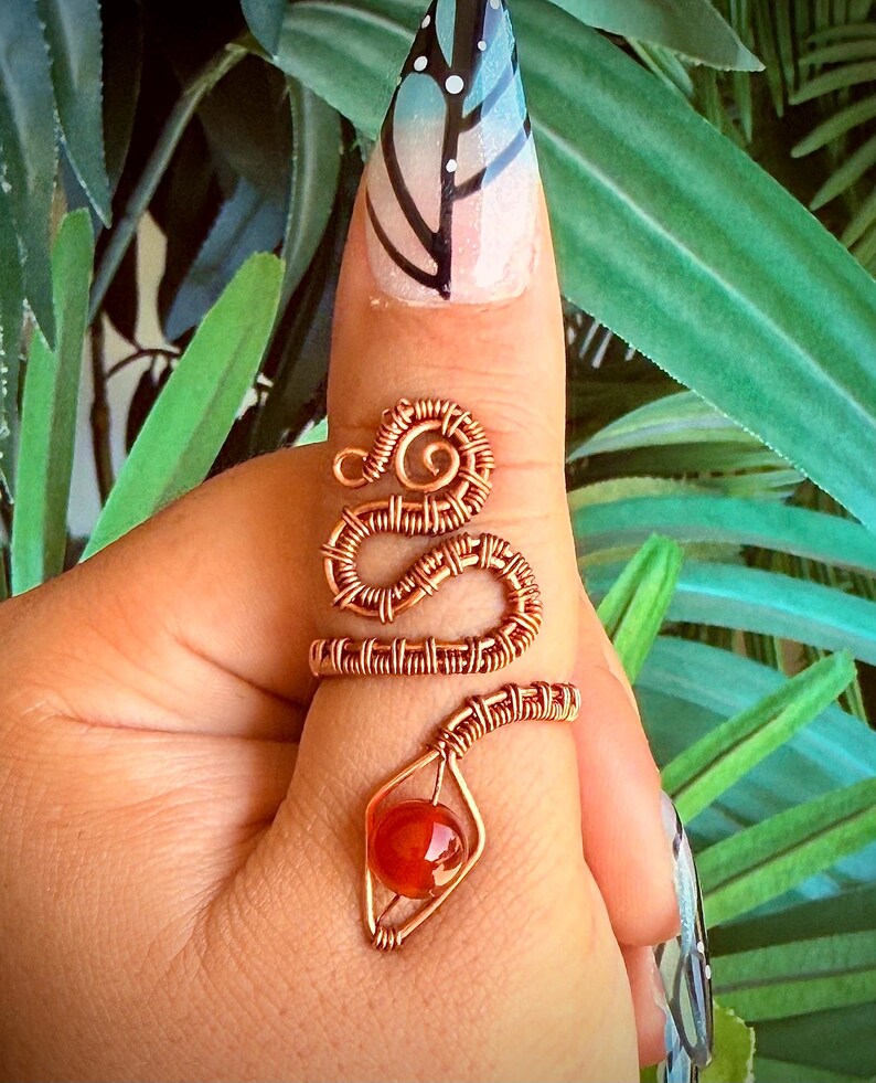 May include: A copper-colored wire-wrapped ring in the shape of a snake, with a red gemstone at the bottom. The ring is worn on a finger with a butterfly nail design. The snake's body coils around the finger.