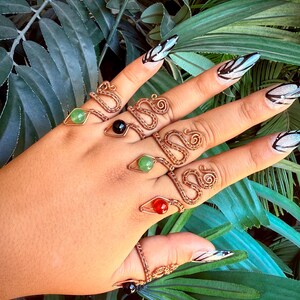 May include: A hand displays several copper wire rings with gemstone accents. The rings feature various designs, including snake-like coils and geometric shapes. Gemstones are green, red, and black. The background includes green foliage.