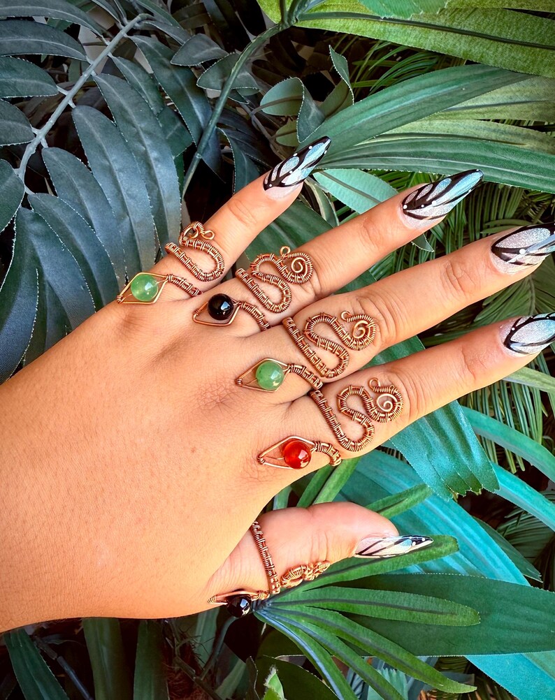 May include: A hand displays several copper wire rings with gemstone accents. The rings feature various designs, including snake-like coils and geometric shapes. Gemstones are green, red, and black. The background includes green foliage.