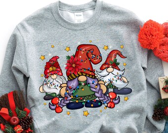 May include: A light gray sweatshirt featuring a festive gnome design. The graphic includes three gnomes with colorful hats, string lights, and star accents. A red knit hat, denim jeans, and a decorative broom are also in the image.