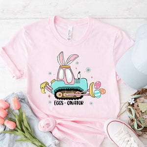 Może przedstawiać: Różowa koszulka z grafiką przedstawiającą kreskówkową koparkę z uszami króliczka i tekstem "Eggs-cavator".