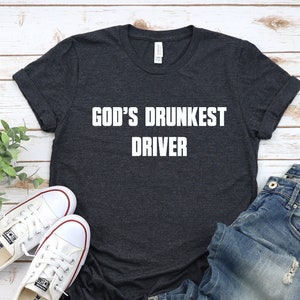 May include: A dark gray t-shirt with the text "GOD'S DRUNKEST DRIVER" in white lettering. The shirt is laid flat on a white wooden surface with a pair of white canvas shoes and blue jeans.