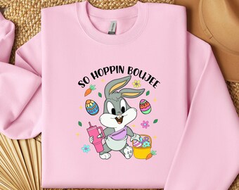 Könnte beinhalten: Rosafarbener Pullover mit einem Cartoon von Bugs Bunny, der einen Korb mit Ostereiern und ein Getränk hält. Der Text "So Hoppin' Boujee" ist auf dem Pullover aufgedruckt.