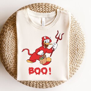 以下が含まれることがあります： 悪魔の格好をしたカモの漫画の画像が描かれた白いTシャツ。カモはフォークとトリックオアトリートバッグを持っています。画像の下には「BOO！」という文字が印刷されています。