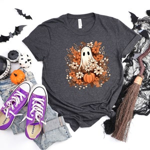 May include: Dark grey t-shirt with a Halloween graphic of a ghost, flowers, and a pumpkin. The image includes purple trainers, jeans, and Halloween decorations. The shirt is made of a soft material.
