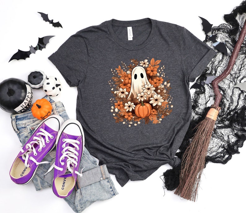May include: Dark grey t-shirt with a Halloween graphic of a ghost, flowers, and a pumpkin. The image includes purple trainers, jeans, and Halloween decorations. The shirt is made of a soft material.