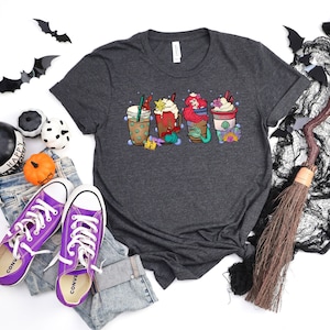 May include: A dark gray t-shirt with a graphic of four coffee cups featuring a mermaid, a starfish, and a seashell. The cups are decorated with red, green, and white accents.