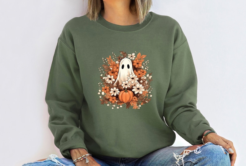 May include: An olive green sweatshirt with a cartoon ghost design. The ghost is white with black eyes, surrounded by orange and white flowers, and a pumpkin. A seasonal design for autumn and Halloween.