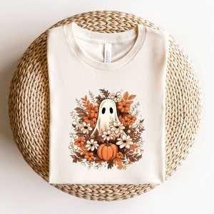 May include: A cream-coloured t-shirt with a Halloween graphic. The design features a friendly ghost surrounded by autumn flowers and a pumpkin. The artwork uses shades of orange, brown, and white, creating a seasonal aesthetic.
