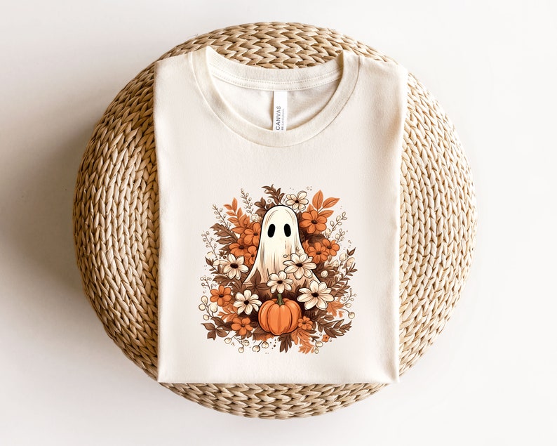 May include: A cream-coloured t-shirt with a Halloween graphic. The design features a friendly ghost surrounded by autumn flowers and a pumpkin. The artwork uses shades of orange, brown, and white, creating a seasonal aesthetic.
