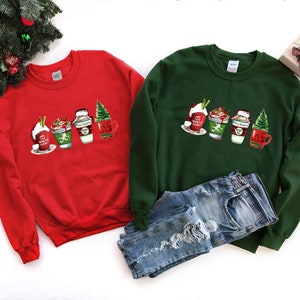 Puede incluir: Dos sudaderas con tem&aacute;tica navide&ntilde;a, una roja y otra verde, con un gr&aacute;fico de cuatro tazas de caf&eacute; con tem&aacute;tica navide&ntilde;a. El texto en las tazas dice "Hot Cocoa", "Let's Get Sippin'" y "Merry Christmas".