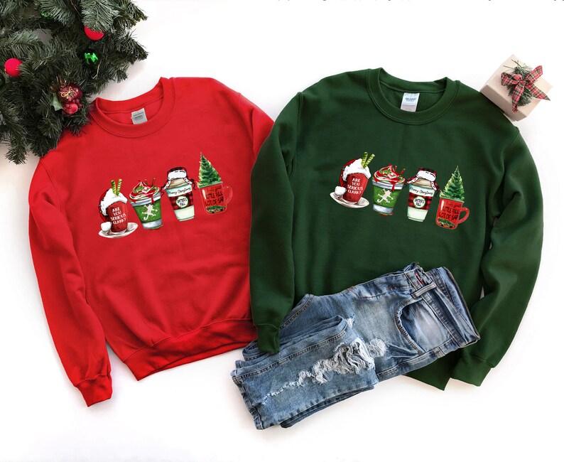 Puede incluir: Dos sudaderas con tem&aacute;tica navide&ntilde;a, una roja y otra verde, con un gr&aacute;fico de cuatro tazas de caf&eacute; con tem&aacute;tica navide&ntilde;a. El texto en las tazas dice "Hot Cocoa", "Let's Get Sippin'" y "Merry Christmas".