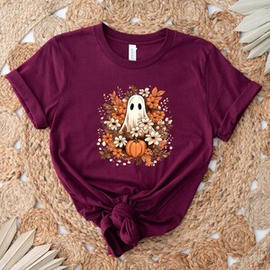 May include: A maroon t-shirt with a ghost design surrounded by autumn flowers and a pumpkin. The ghost has a friendly expression. The design is centered on the shirt, which is folded at the bottom.