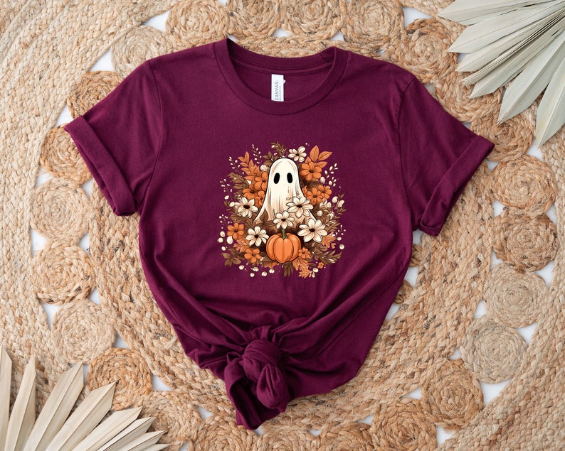 May include: A maroon t-shirt with a ghost design surrounded by autumn flowers and a pumpkin. The ghost has a friendly expression. The design is centered on the shirt, which is folded at the bottom.