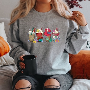 May include: A gray sweatshirt with a graphic of four different coffee drinks. The drinks are decorated with whipped cream, sprinkles, and other toppings. The sweatshirt is being worn by a person sitting on a couch.