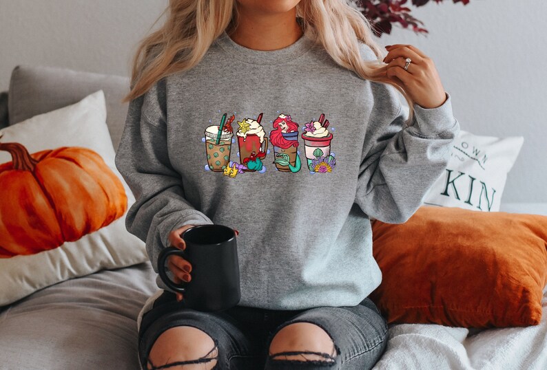 May include: A gray sweatshirt with a graphic of four different coffee drinks. The drinks are decorated with whipped cream, sprinkles, and other toppings. The sweatshirt is being worn by a person sitting on a couch.