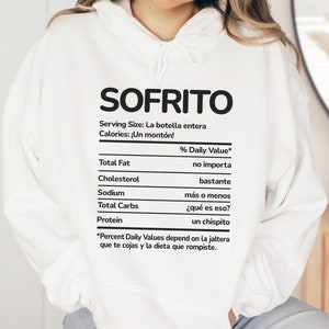 May include: White hoodie with the word "SOFRITO" in large black letters. Below, a nutrition facts label humorously lists "no importa" for fat, "bastante" for cholesterol, and "un chispito" for protein. The hoodie has a drawstring hood.