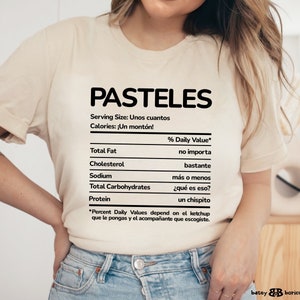 May include: A white t-shirt with a nutrition facts label that says "PASTELES" at the top. The label lists "Serving Size: Unos cuantos" and "Calories: &iexcl;Un mont&oacute;n!"  The label also lists the % Daily Value for Total Fat, Cholesterol, Sodium, Total Carbohydrates, and Protein. The label is written in Spanish.