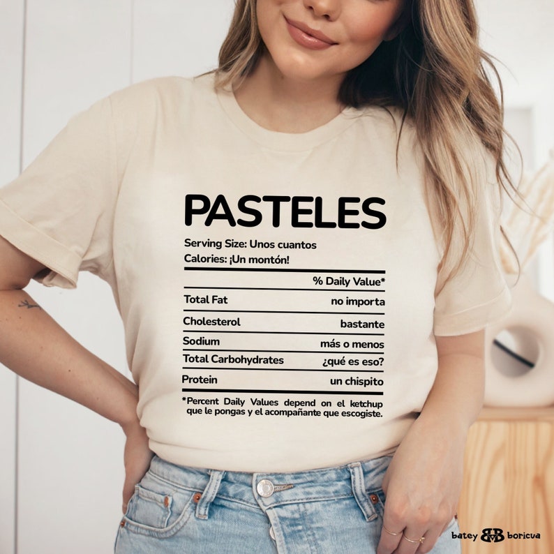 May include: A white t-shirt with a nutrition facts label that says "PASTELES" at the top. The label lists "Serving Size: Unos cuantos" and "Calories: &iexcl;Un mont&oacute;n!"  The label also lists the % Daily Value for Total Fat, Cholesterol, Sodium, Total Carbohydrates, and Protein. The label is written in Spanish.