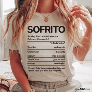 May include: White t-shirt with the word "SOFRITO" in large black letters. Below, a nutrition facts label humorously lists "Total Fat," "Cholesterol," "Sodium," "Total Carbs," and "Protein" with funny responses. The shirt's theme is food.