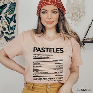 May include: A pink t-shirt with a nutrition facts label that says "PASTELES" with the text "Serving Size: Unos cuantos" and "Calories: &iexcl;Un mont&oacute;n!"  The label lists the nutritional information for the food, with humorous descriptions for each category.  The text on the label is in Spanish.