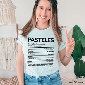 May include: A light blue t-shirt with a nutrition facts label that says "PASTELES" and has a humorous take on the nutritional information. The label says "Serving Size: Unos cuantos" and "Calories: &iexcl;Un mont&oacute;n!"  The % Daily Value for Total Fat is "no importa", Cholesterol is "bastante", Sodium is "m&aacute;s o menos", Total Carbohydrates is "&iquest;qu&eacute; es eso?", and Protein is "un chispito".