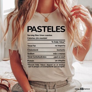 May include: White t-shirt with a black graphic of a nutrition facts label that says "PASTELES" at the top. The label says "Serving Size: Unos cuantos" and "Calories: &iexcl;Un mont&oacute;n!" The label lists "Total Fat", "Cholesterol", "Sodium", "Total Carbohydrates", and "Protein" with humorous Spanish phrases for the % Daily Value.