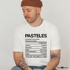 May include: A white t-shirt with a black graphic of a nutrition facts label for "PASTELES". The label says "Serving Size: Unos cuantos" and "Calories: &iexcl;Un mont&oacute;n!" with a disclaimer at the bottom that says "*Percent Daily Values depend on el ketchup que le pongas y el acompa&ntilde;ante que escogiste."